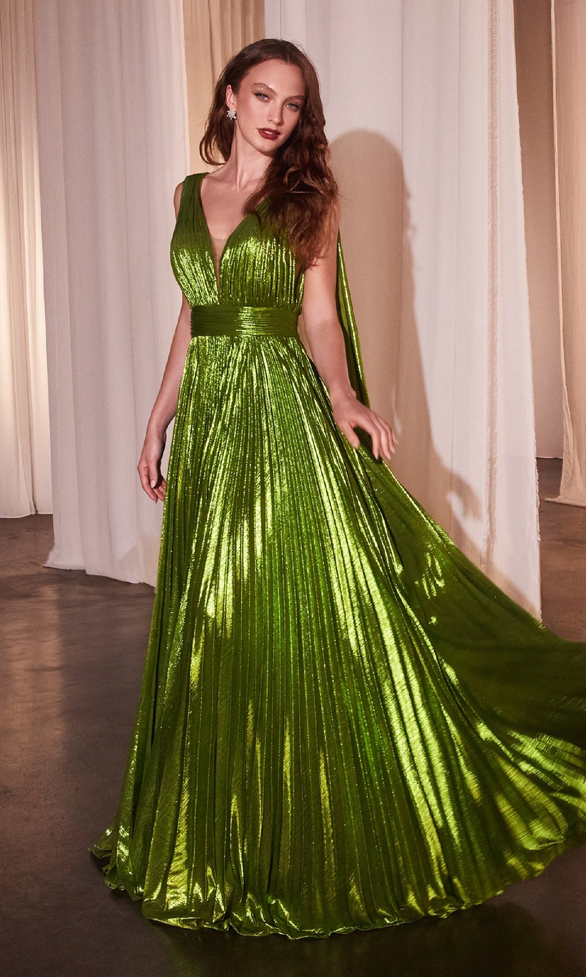 A woman wears the Ladivine CD802 long prom dress, featuring a metallic pleated A-line skirt, sleeveless V-neck bodice, wide waistband, and cape detail, standing before draped white curtains.